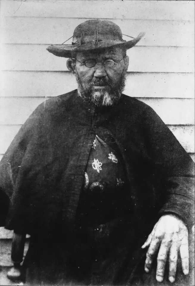 Black-and-white photograph of Father Damien seated in a dark cassock and a straw hat, his face and hand marked by Hansen's disease, wearing glasses