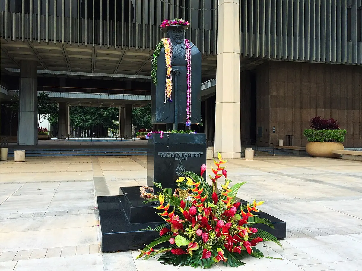 A dark bronze statue of a cloaked figure in a broad hat, hung with lei, on a black stone base surrounded by red and yellow tropical flowers, in front of the Hawaii State Capitol
