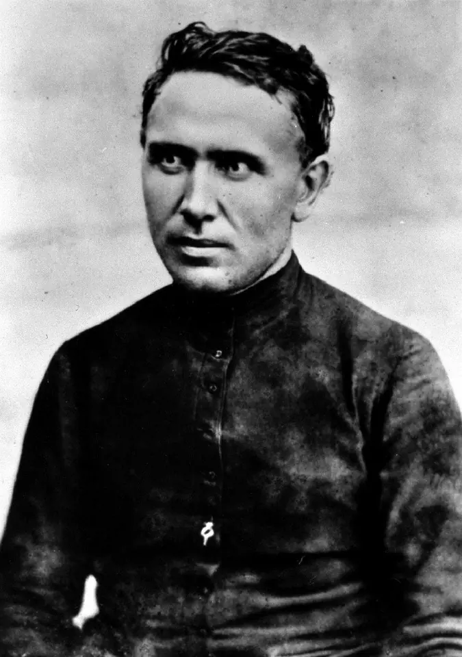 Black-and-white photograph of a young, clean-shaven Father Damien with thick dark hair, in a black cassock, looking to the side