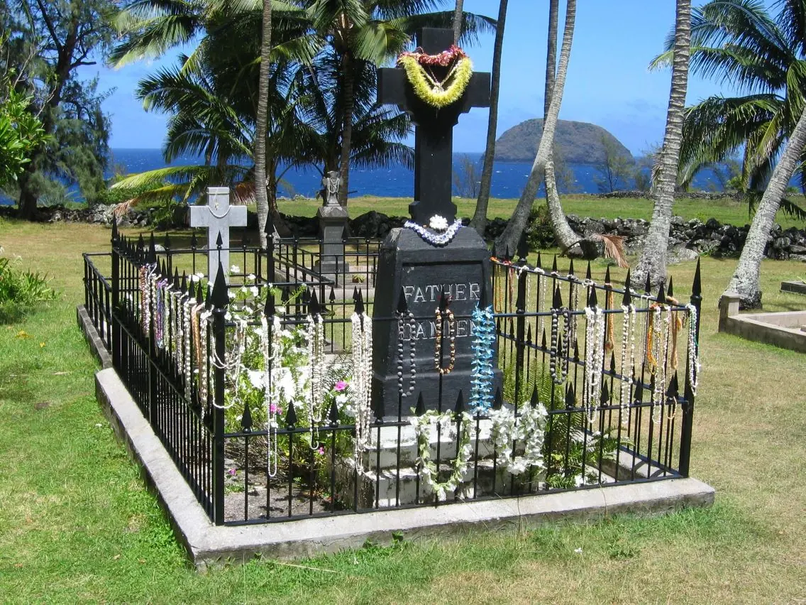A black stone cross inside an iron fence hung with flower lei, on green grass, with palm trees, the ocean and a small island behind