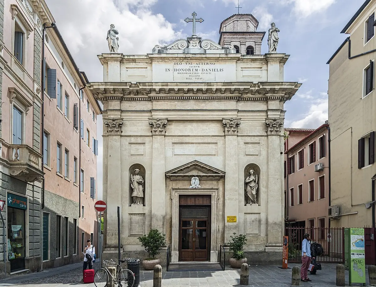 Pale stone church front with columns, a triangular pediment, statues in two niches and a carved dedication, on a narrow street