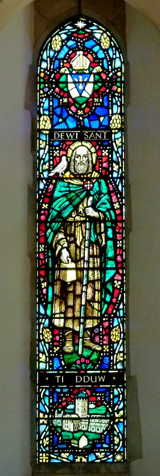 Tall narrow stained glass window labeled "Dewi Sant" showing the saint in green and white robes, with a dove and a small church below