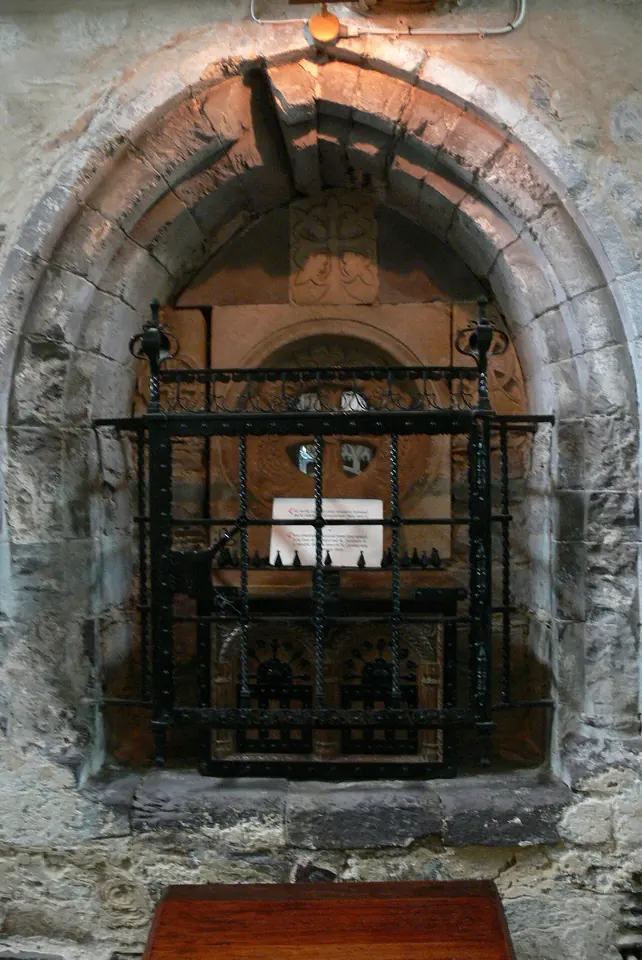 A wooden casket behind an iron grille in a recess of a stone wall, under a rounded arch, in a dimly lit chapel