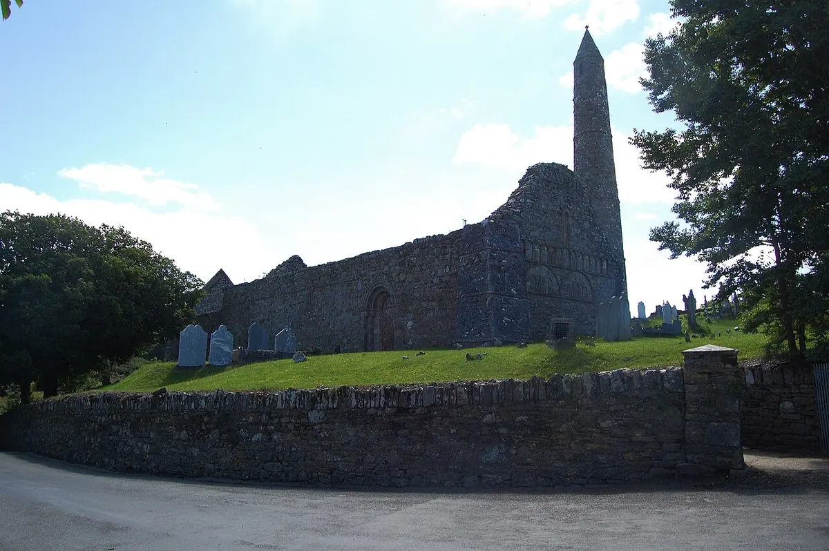 Roofless medieval stone church beside a tall slender round tower with a conical cap, above a graveyard wall under a bright sky