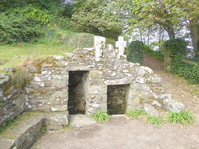 Low rough stone well-house with two dark openings and two small stone crosses on top, set among ferns and trees