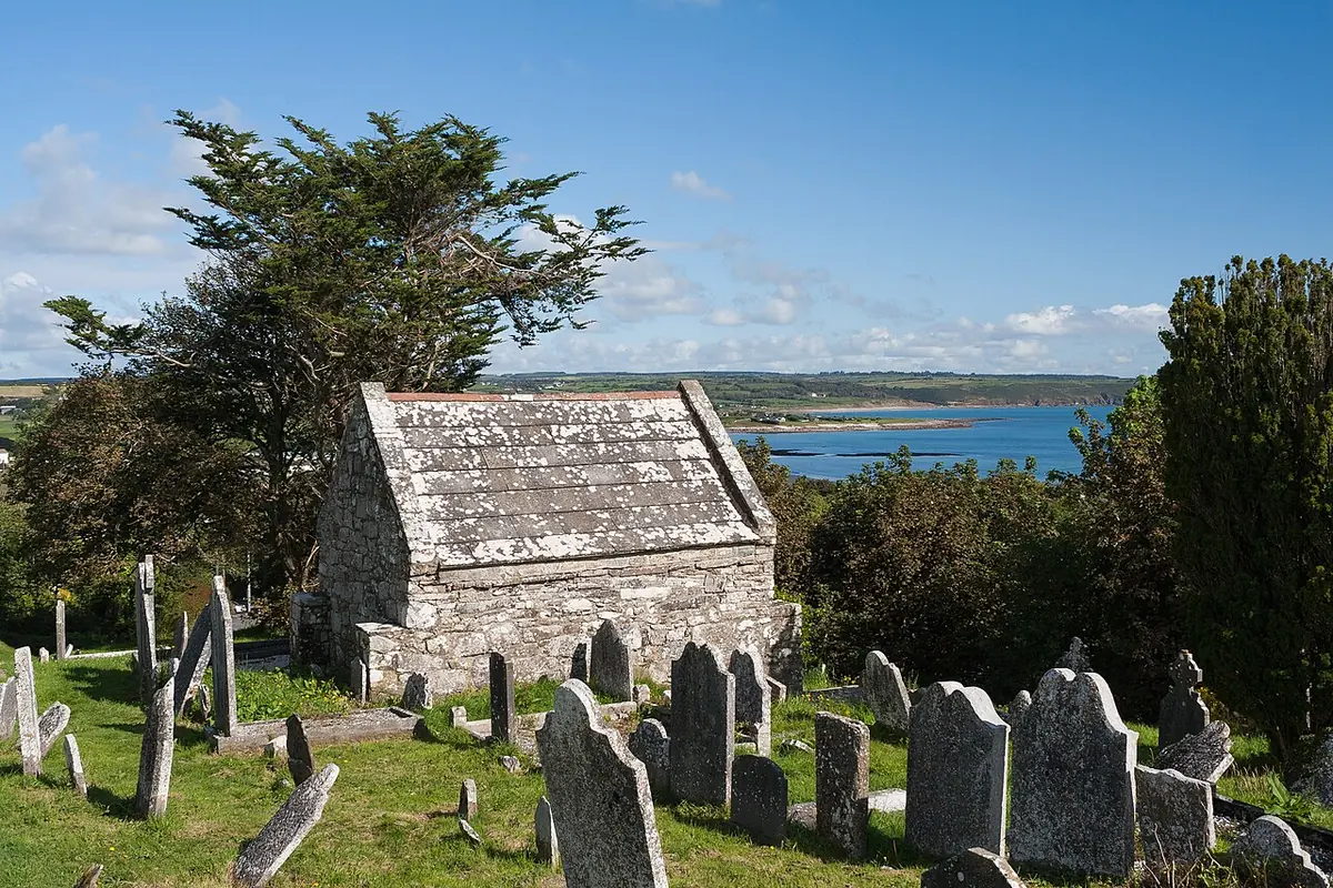 Small rectangular stone oratory with a steep slate roof among old gravestones, with the sea and a distant headland beyond