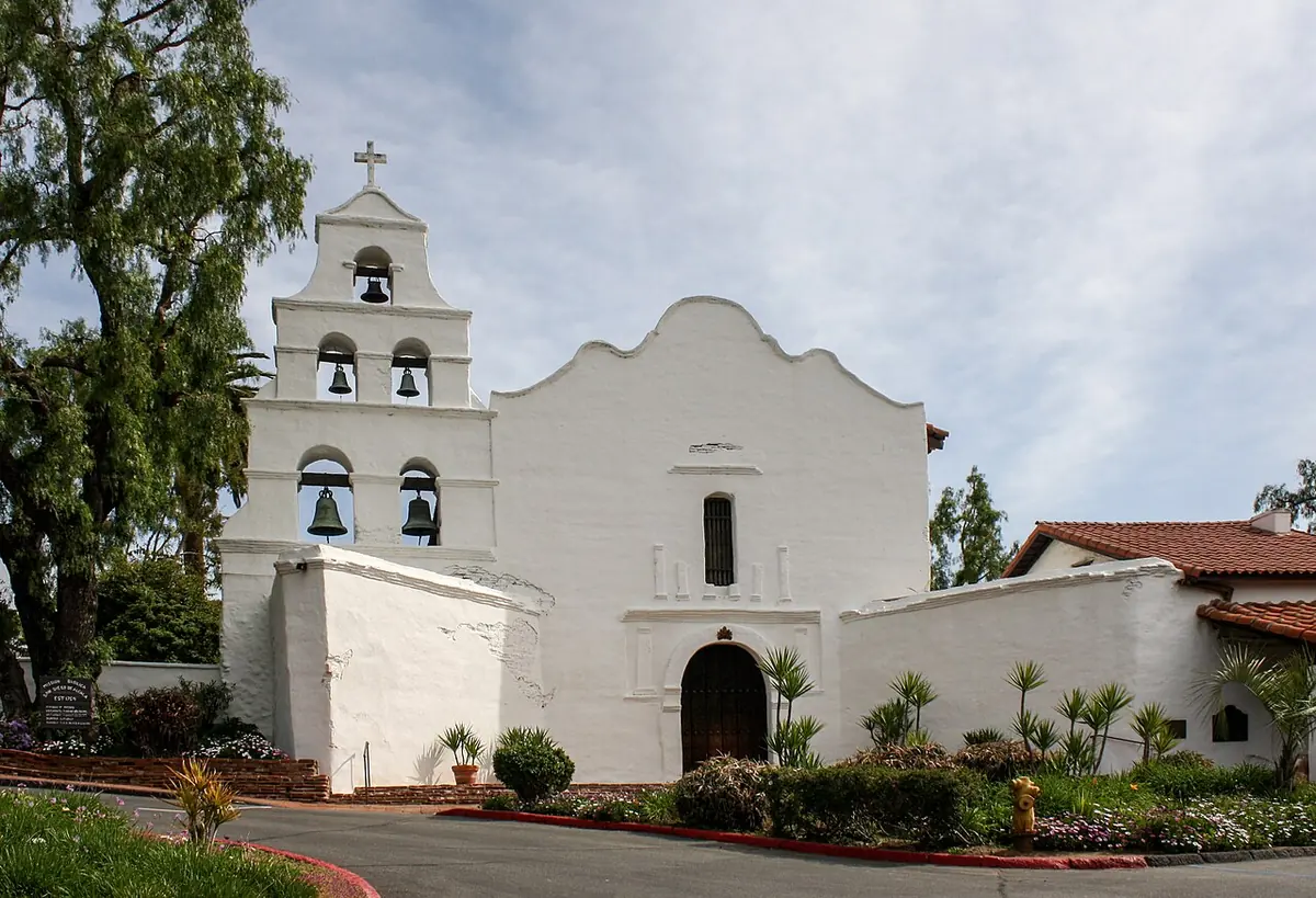 White adobe mission church with a tall bell wall holding five bells under a cross, a palm-lined garden and a low tiled building beside it