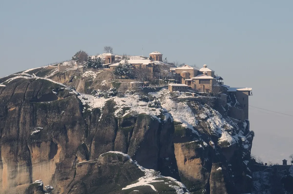 A monastery with tiled roofs perched on top of a sheer rock pillar dusted with snow