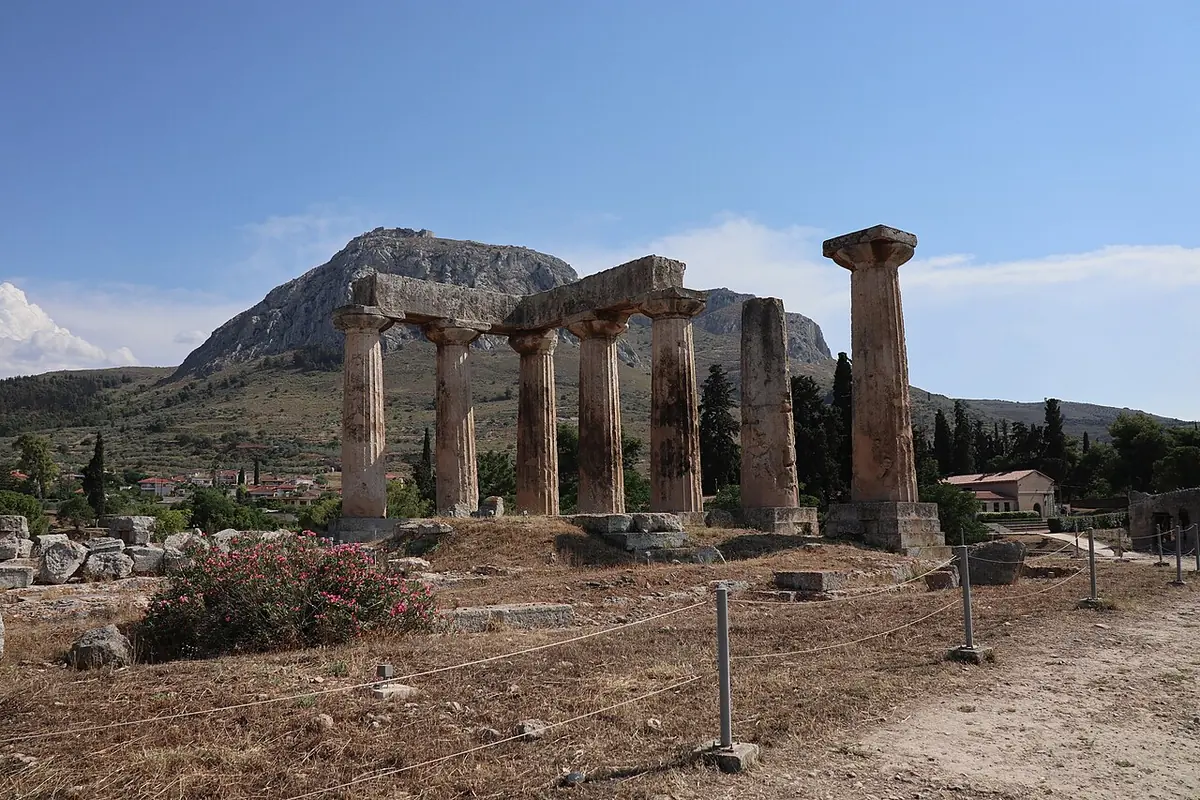 The seven standing Doric columns of the Temple of Apollo at ancient Corinth, with the rock of Acrocorinth rising behind them under a blue sky
