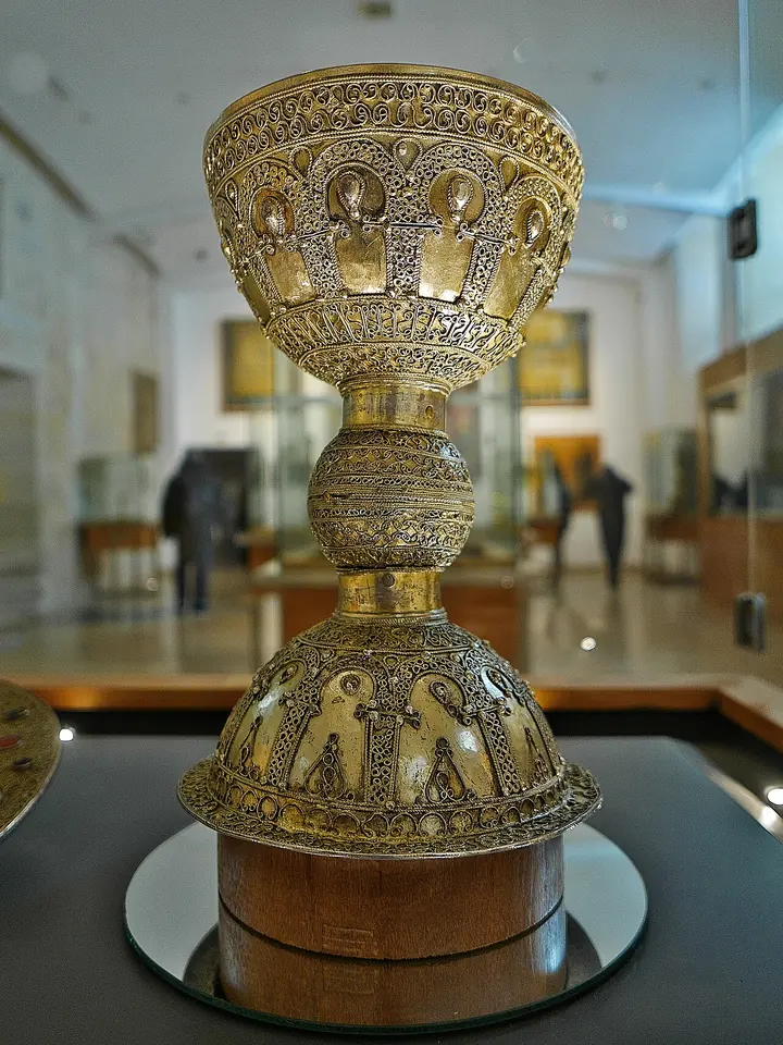 Tall gilded silver chalice with a wide cup and a domed foot covered in filigree and small arched panels, in a museum case