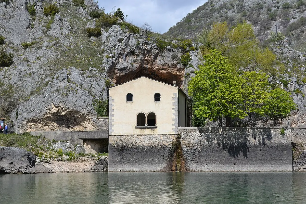 A small whitewashed hermitage church built against a rocky cliff at the edge of a green mountain lake