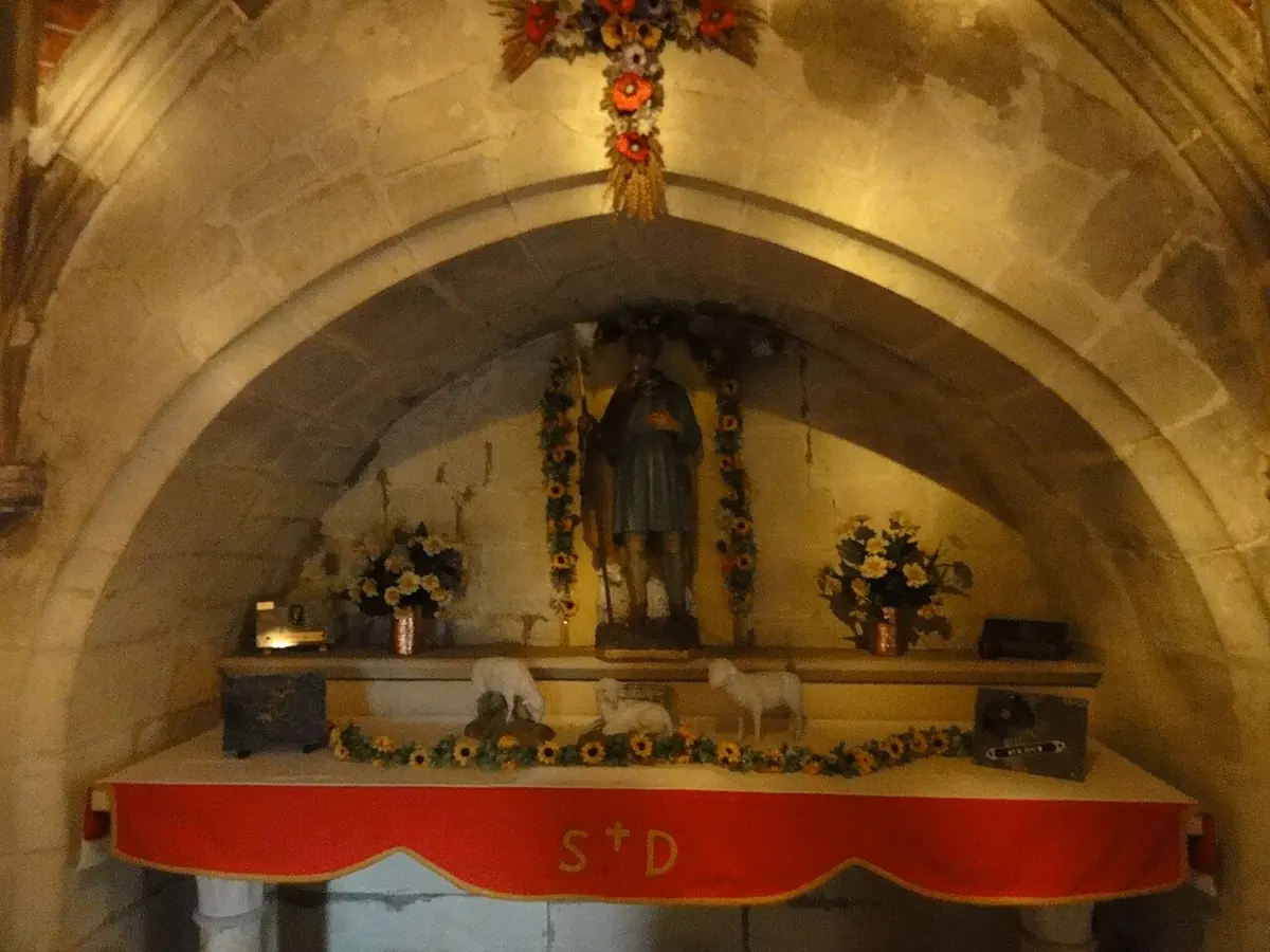 A small side altar under a stone arch with a statue of the shepherd saint in a niche, framed by flowers and small white sheep figures, with a red altar cloth marked St D