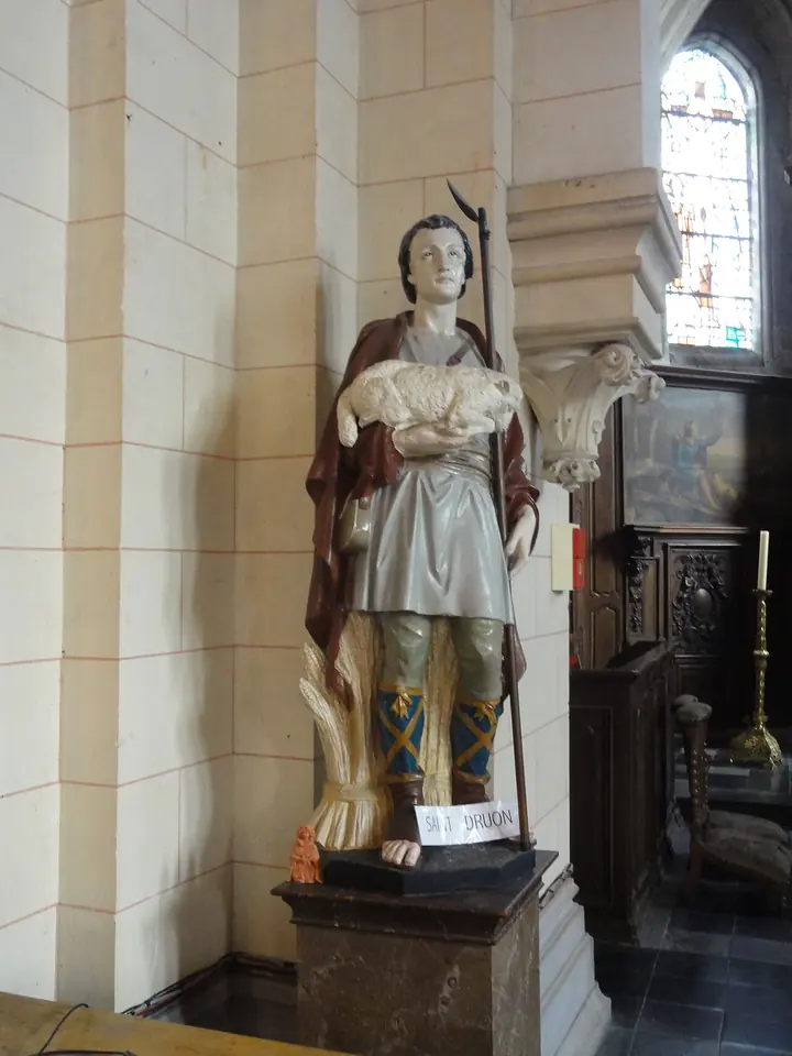 Painted wooden statue of a young shepherd in a red cloak and gray tunic carrying a white lamb in his arms, standing beside a pillar in a church