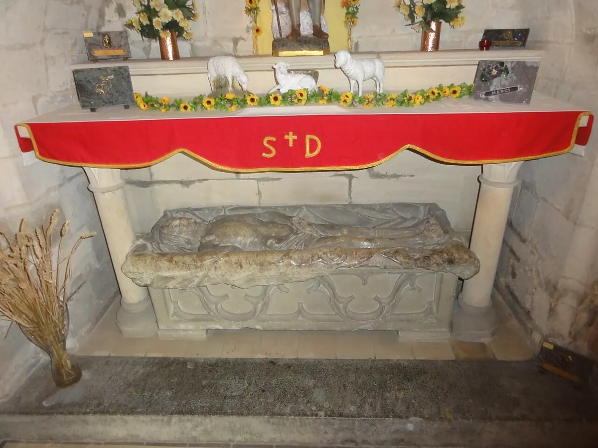 Under a stone altar with a red cloth marked St D lies an old stone tomb with a worn carved figure, with a sheaf of wheat leaning against it