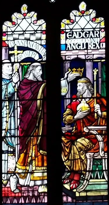 Stained-glass window of an archbishop in a purple robe placing a crown on the head of a seated king who holds an orb, with the words Eadgar Angl Rex above
