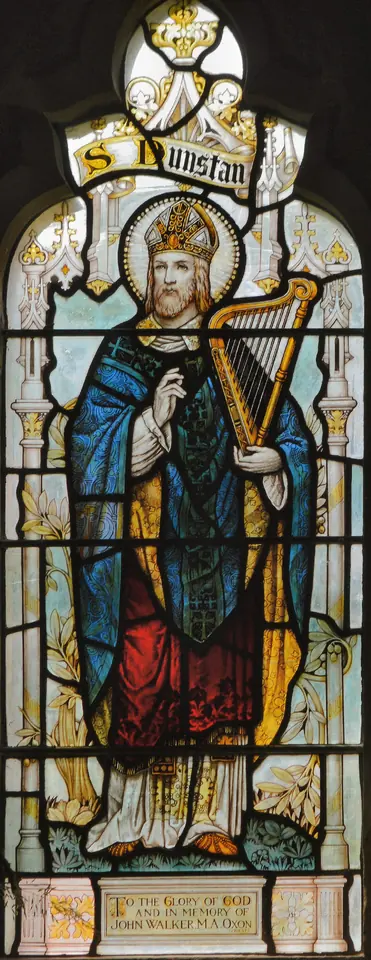 Stained-glass window of St. Dunstan standing in a blue cloak and miter, holding a golden harp, with his name above