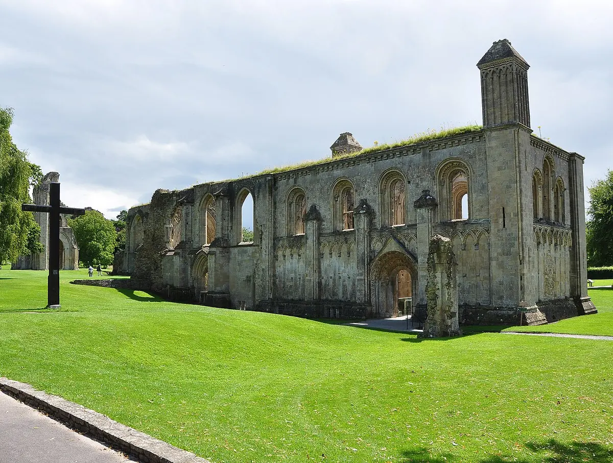 The roofless stone walls of a medieval chapel at Glastonbury Abbey, with tall arched windows, on a green lawn under a cloudy sky