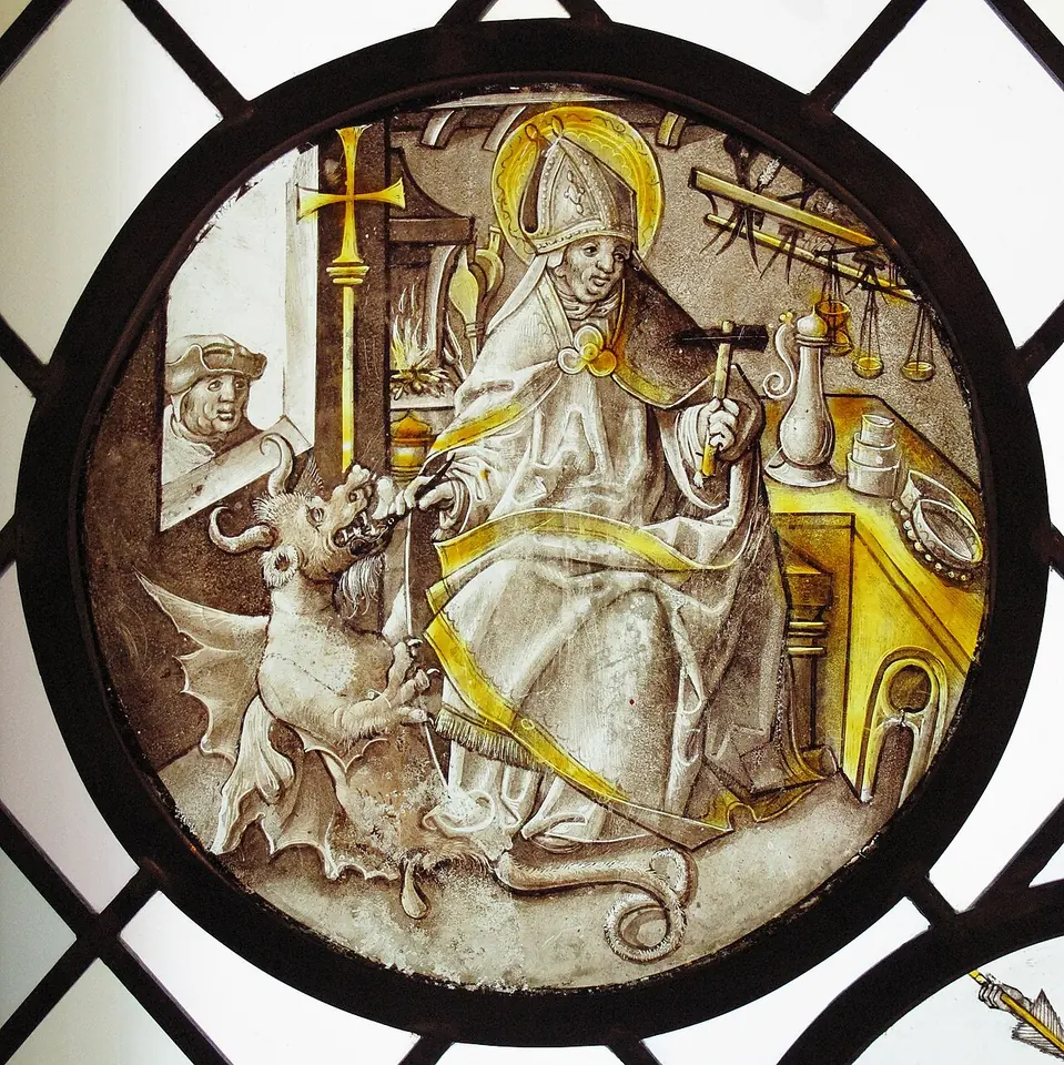 Round stained-glass panel of St. Dunstan seated at a workbench in miter and vestments, gripping a horned, winged devil by the nose with tongs, with goldsmith's tools and a chalice around him