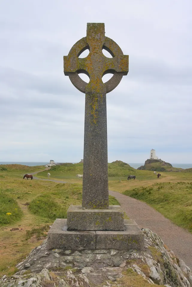 A tall weathered stone Celtic cross on a rocky outcrop above a grassy path, with ponies grazing and a white lighthouse in the distance