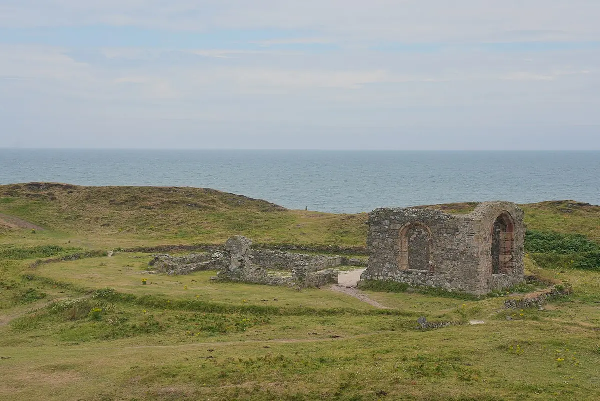The roofless stone ruins of a small church with an empty arched window on a green, grassy island, with the sea and a pale sky beyond