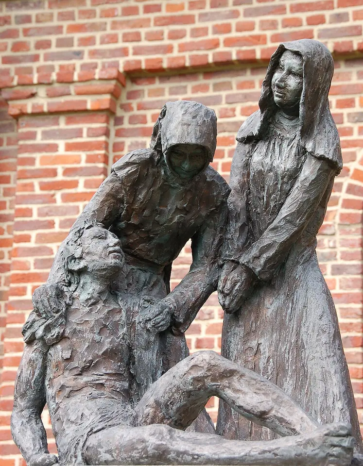 Bronze sculpture of two women in plain hooded dresses bending to lift and support a weak, half-seated man, against a brick wall