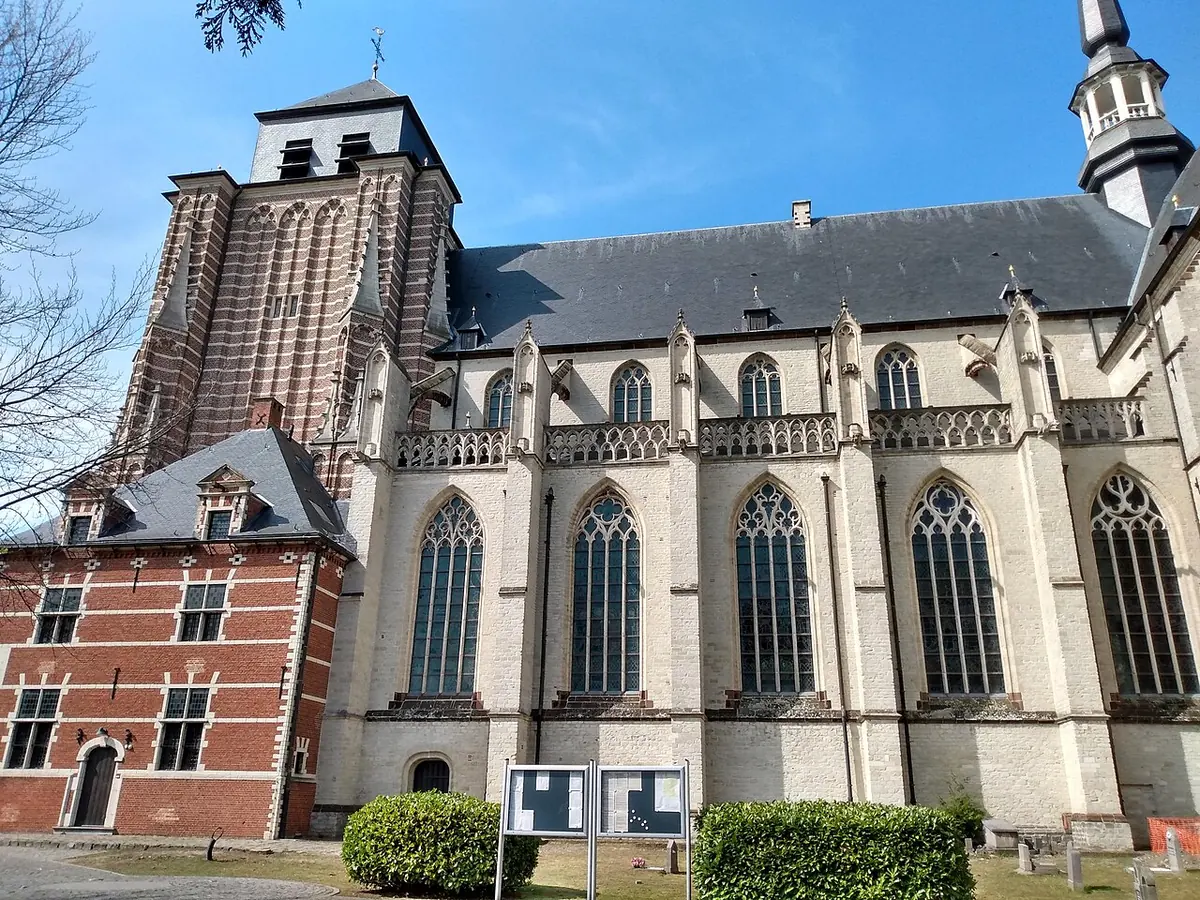 The long Gothic side of the Church of St. Dymphna in pale stone and red brick, with tall pointed windows and a slender spire, behind a lawn