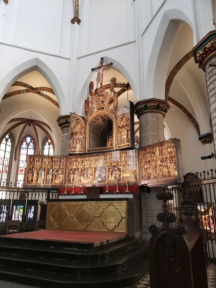 A long gilded carved wooden altarpiece crowded with small figures in Gothic niches stands behind the high altar in a white vaulted choir