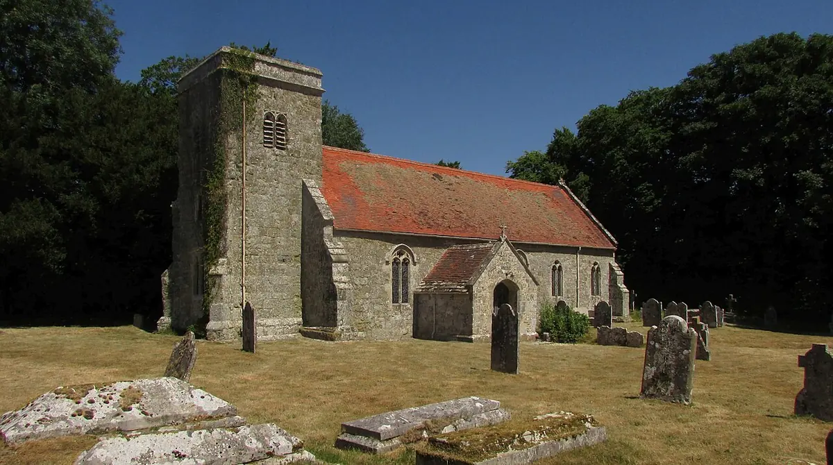 Small medieval flint and stone church with a square tower and red-tiled roof in a churchyard with old gravestones under a blue sky