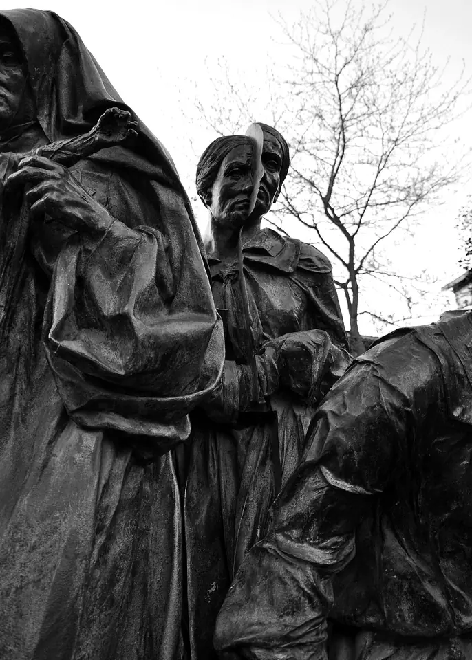 Black-and-white photo of a bronze sculpture group of robed and cloaked figures beside a bare winter tree