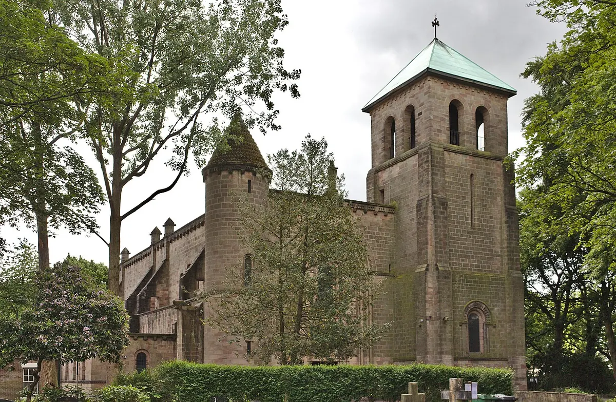 A sandstone church with a square bell tower topped by a green copper roof, surrounded by tall trees and a trimmed hedge