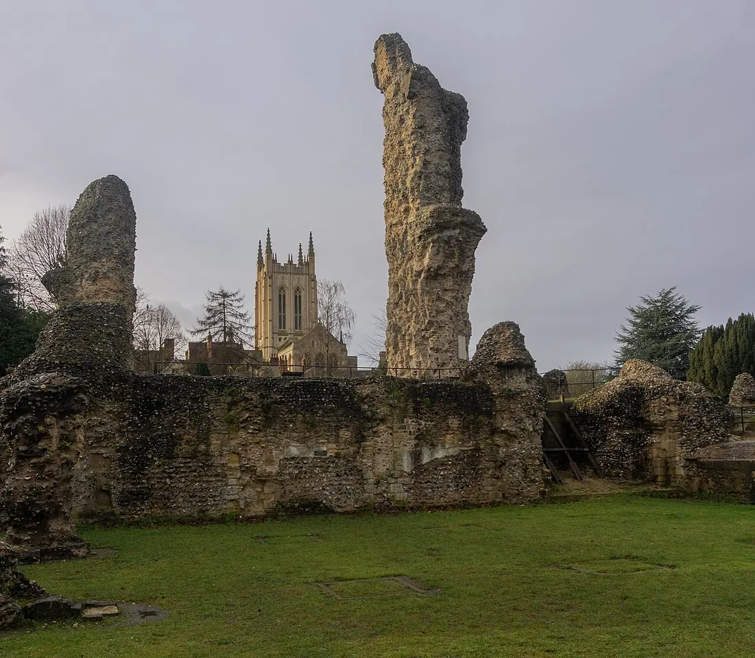 Rough flint ruins of the medieval abbey church standing on a lawn, with the square stone tower of St Edmundsbury Cathedral behind them under a gray sky