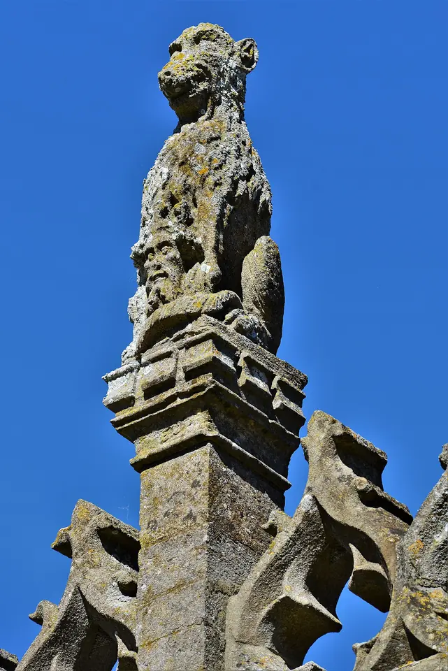 A weathered stone carving of a seated wolf holding a small crowned head between its paws, on top of a church porch pinnacle against a blue sky