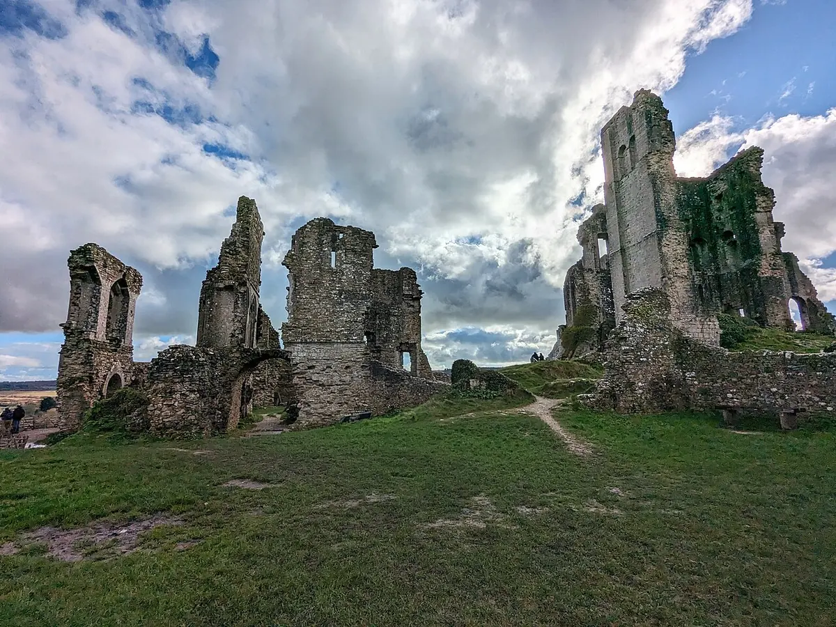 The broken stone walls and towers of Corfe Castle on a grassy hilltop under a cloudy sky