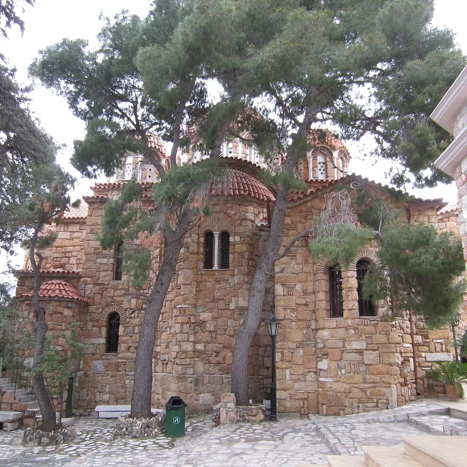 An old Byzantine-style stone church with red-tiled domes, shaded by tall pine trees in a paved courtyard