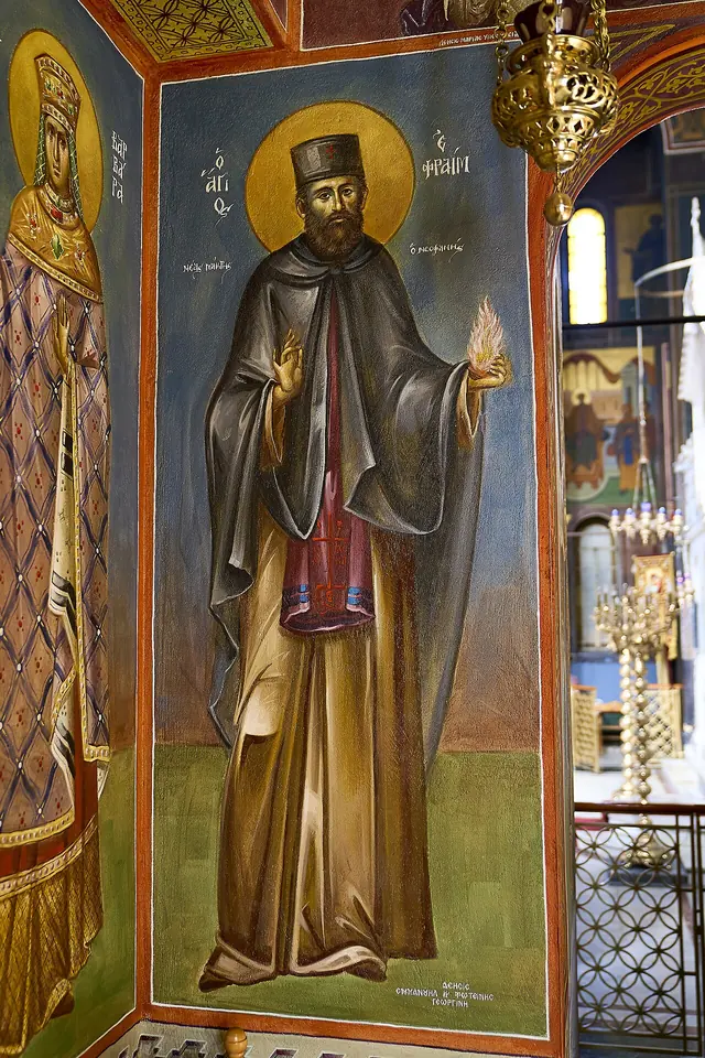 Brightly painted wall icon of St. Efraim in a dark monastic robe holding a cross, in a richly decorated church chapel