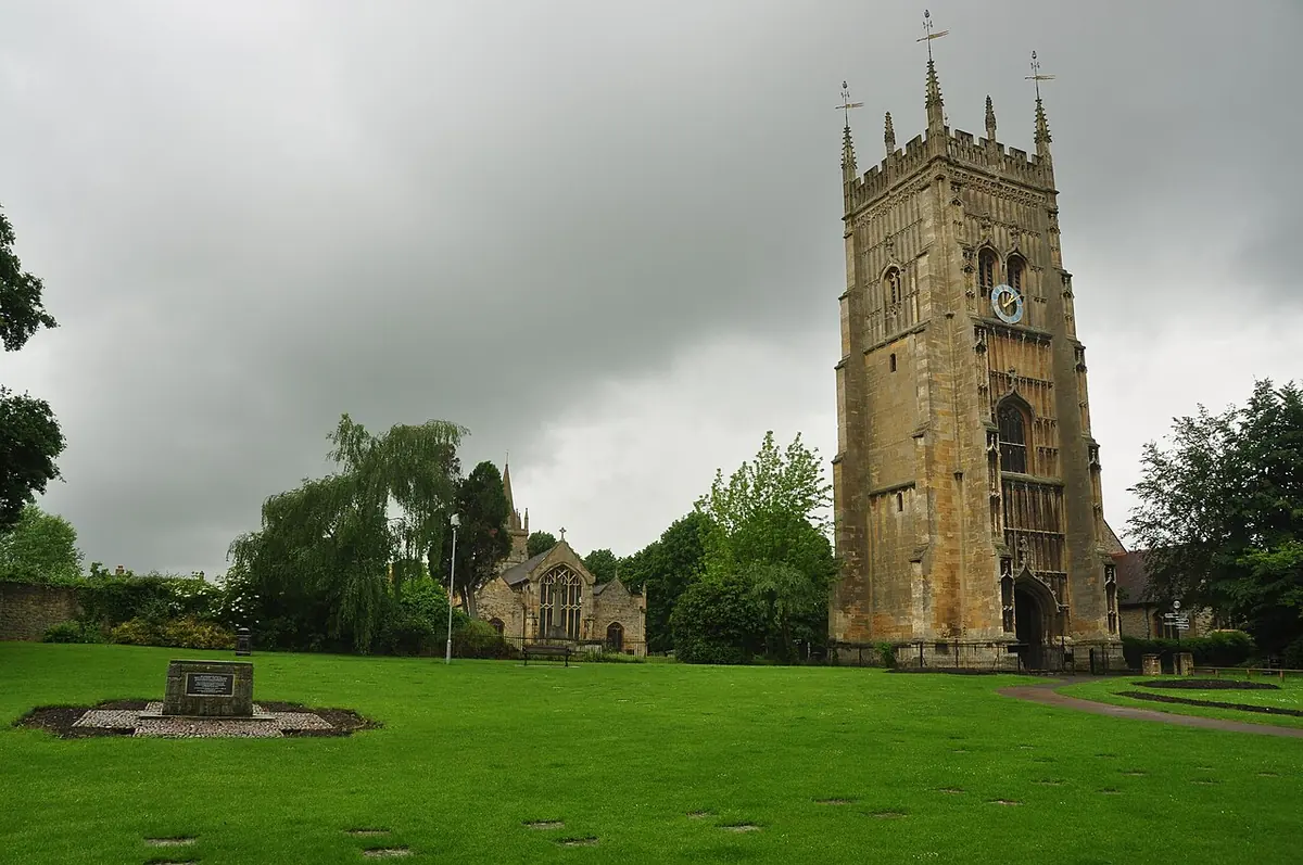 A tall, richly paneled Gothic stone bell tower with pinnacles standing in a broad green lawn, with trees and an old church behind it