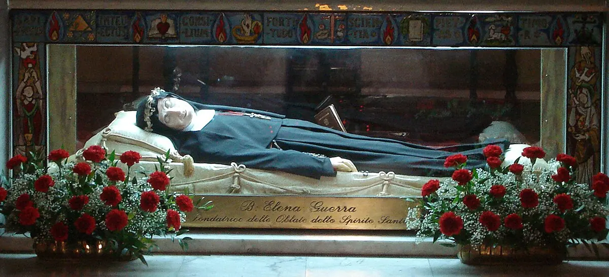 A long glass and gilt urn lined with white satin holding the figure of a nun in a black habit, with red roses at both ends