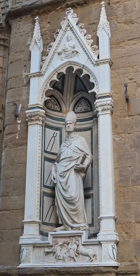 A marble statue of a bearded bishop in miter and flowing robes standing in a white Gothic niche on the wall of Orsanmichele, with a small carved relief below