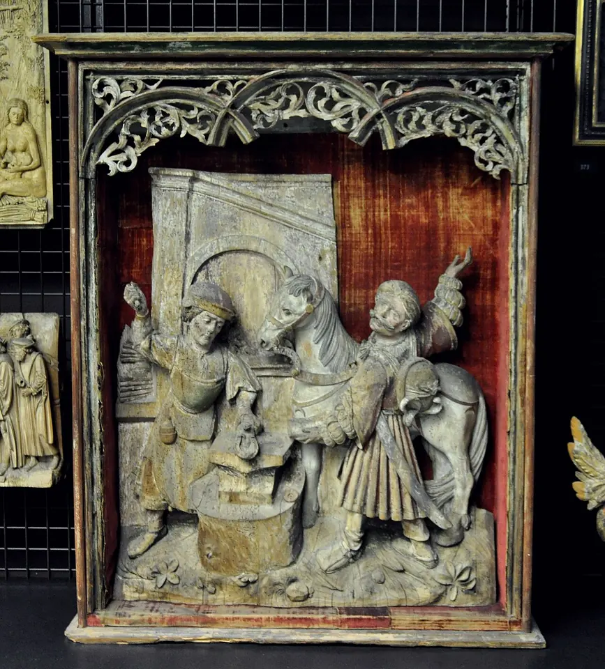 Carved wooden relief in a shrine-like case showing a smith working at an anvil beside a horse held by a young man in a feathered cap