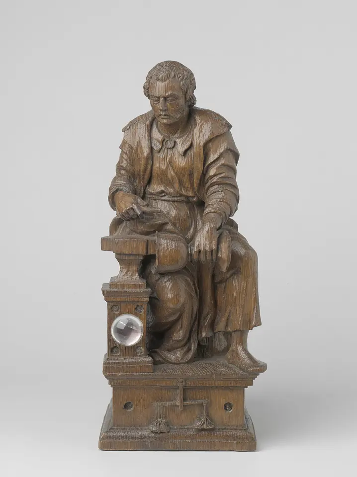Small carved wooden statuette of a seated man in a workman's smock hammering a cup on an anvil set on a tall pedestal