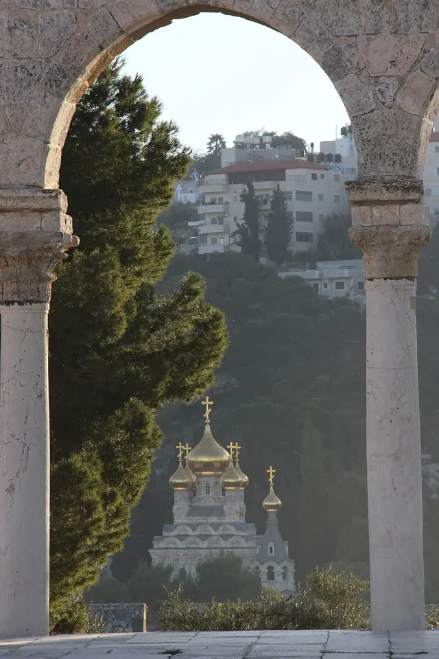 Golden onion domes of a Russian church among pine trees on a hillside, framed by a stone arch