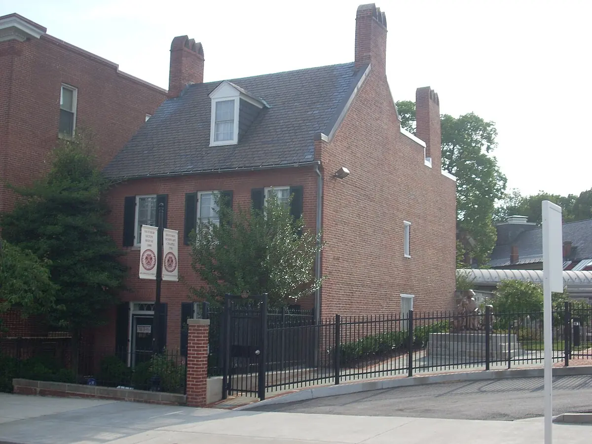 A small two-story red brick Federal house with dormer windows and tall chimneys behind an iron fence