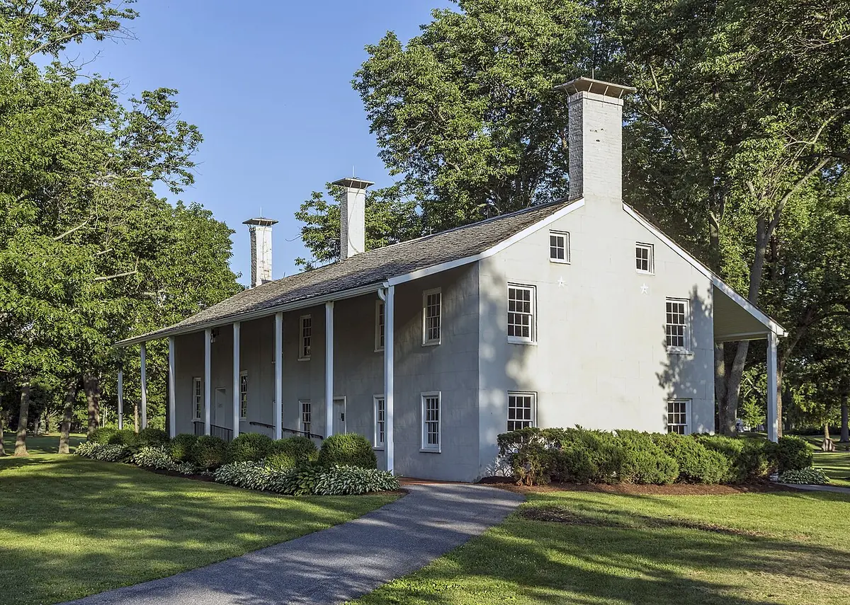 A long white-plastered farmhouse with a deep front porch and two chimneys, set on a lawn under large trees