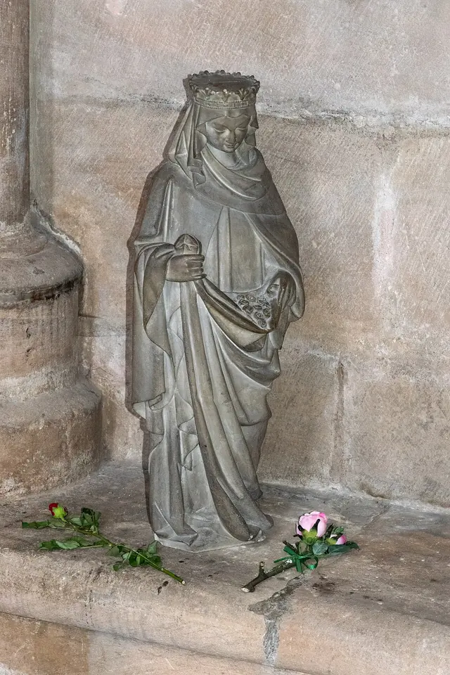 Gray stone Gothic statue of Elizabeth in a crown and long veil holding a small object, with fresh roses laid at its foot by visitors
