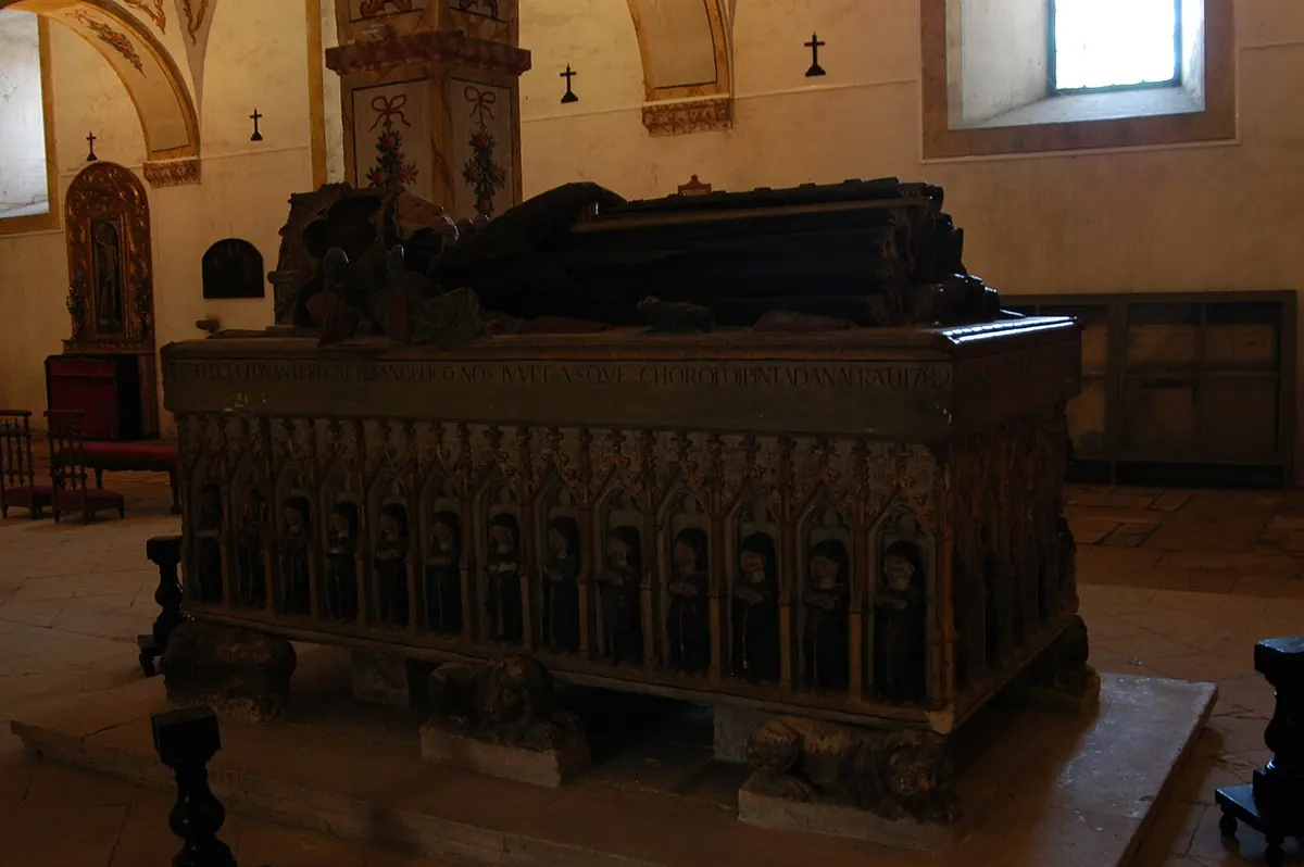 A carved stone Gothic sarcophagus with rows of small figures along its sides and a recumbent effigy on the lid, in a dim church interior
