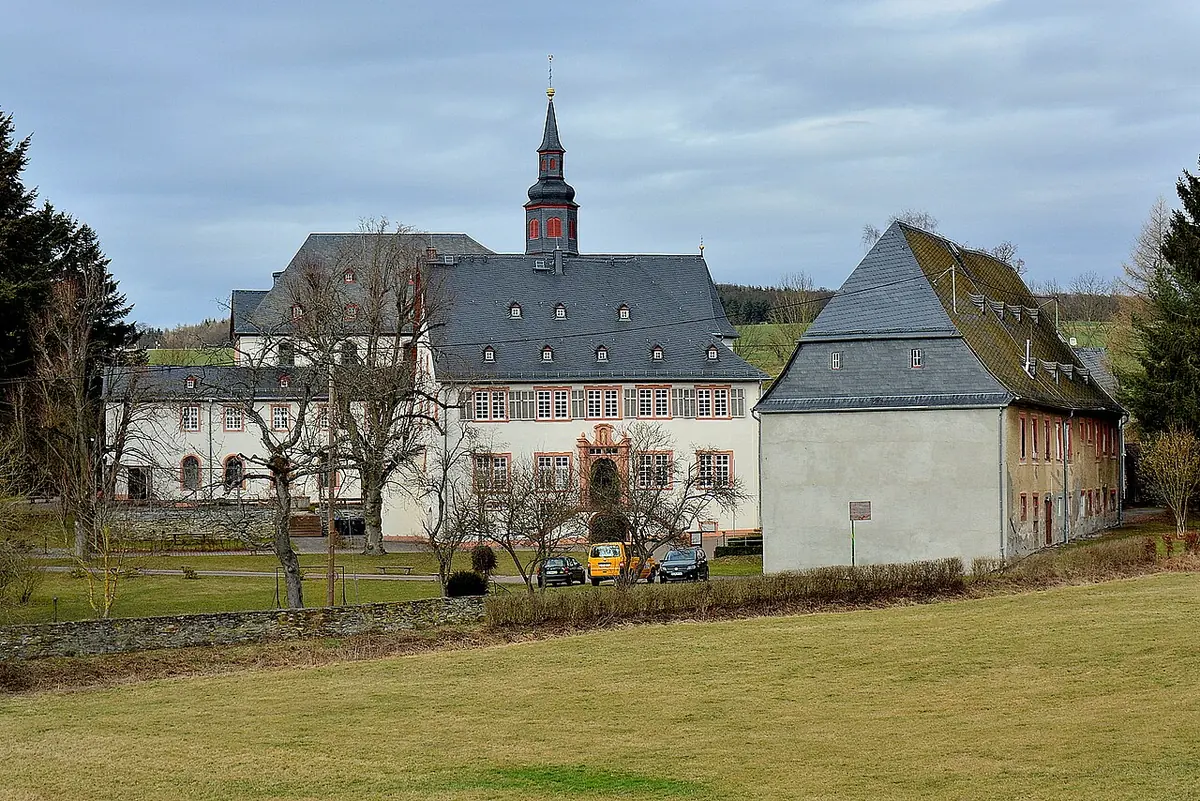 A long white baroque monastery building with a slate roof and a slender church tower, seen across a meadow under a gray sky