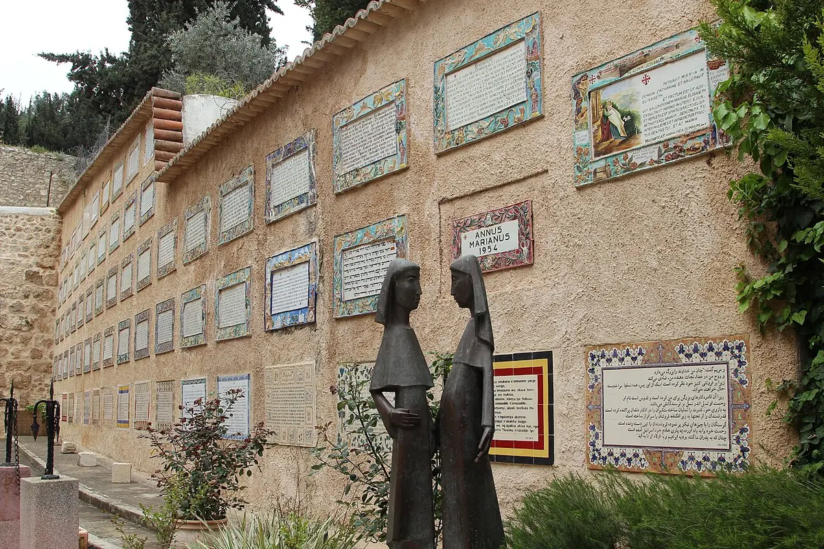 A bronze statue of two slender veiled women facing each other in a courtyard, in front of a wall covered with ceramic tile panels of the Magnificat in many languages
