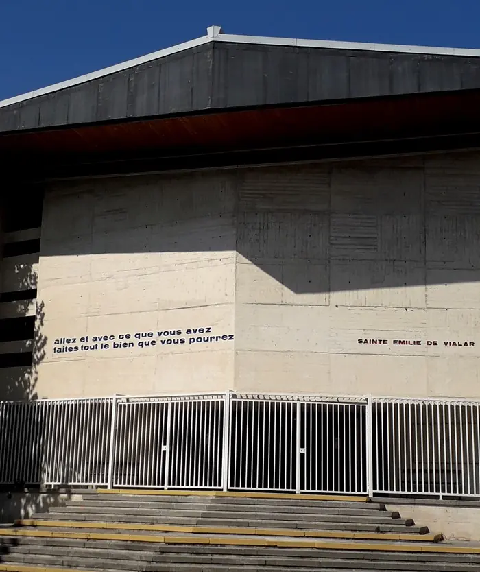 The bare concrete facade of a modern church above a flight of steps, with a line of French words painted across it
