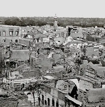 An old black-and-white photograph looking over a district of roofless, ruined stone houses, with a minaret and trees in the distance
