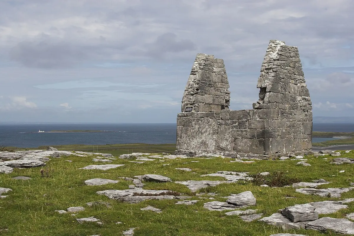 A very small roofless stone church with two tall pointed gables standing on bare limestone rock above the sea
